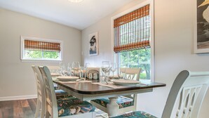 Een eetgedeelte met een houten tafel gedekt voor vier personen, witte stoelen met bloemenmotieven en twee ramen met zonwering.