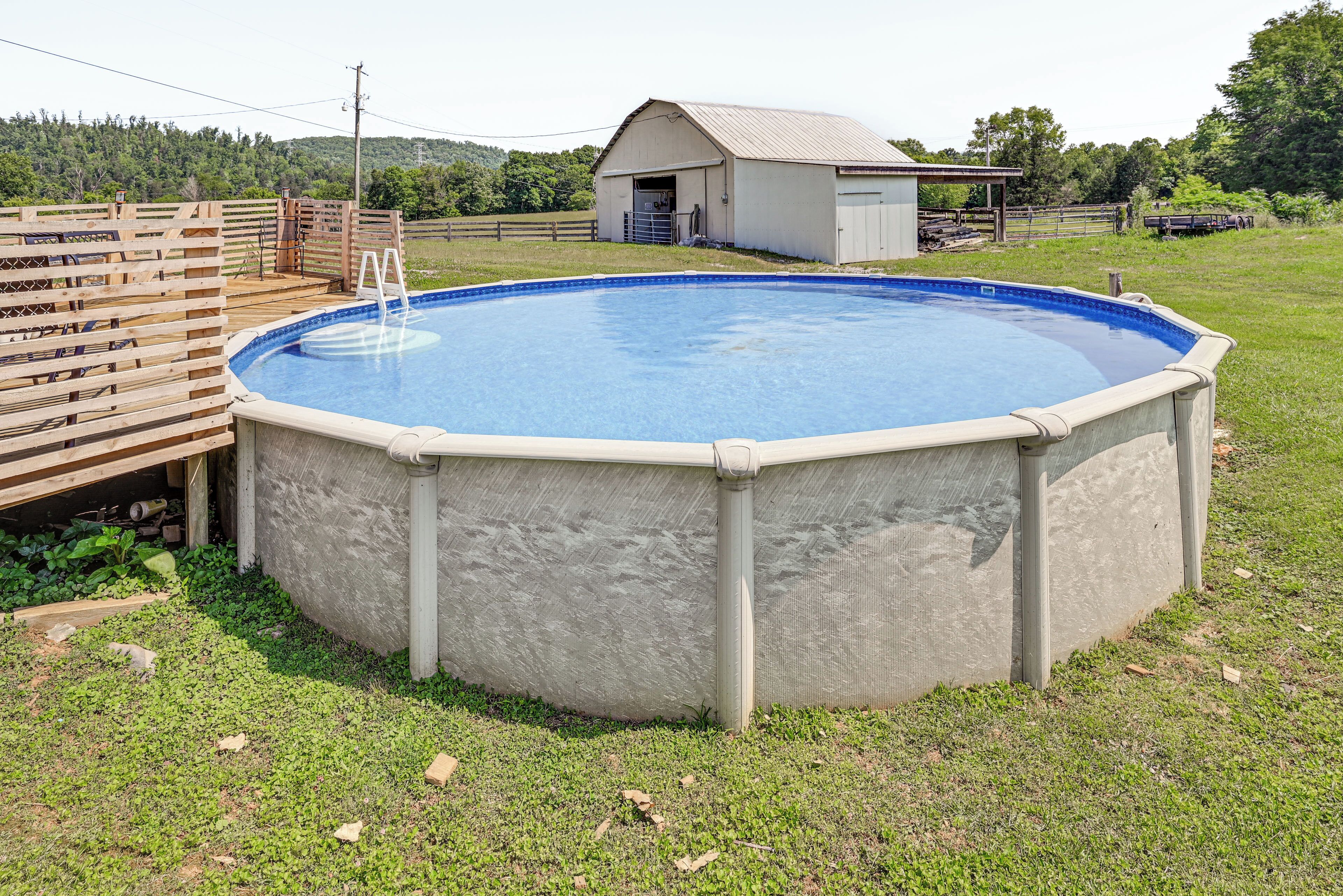 Ein runder, aufgestellter Swimmingpool mit Holzterrasse und einer Leiter, umgeben von einem Rasen und einem Stall.