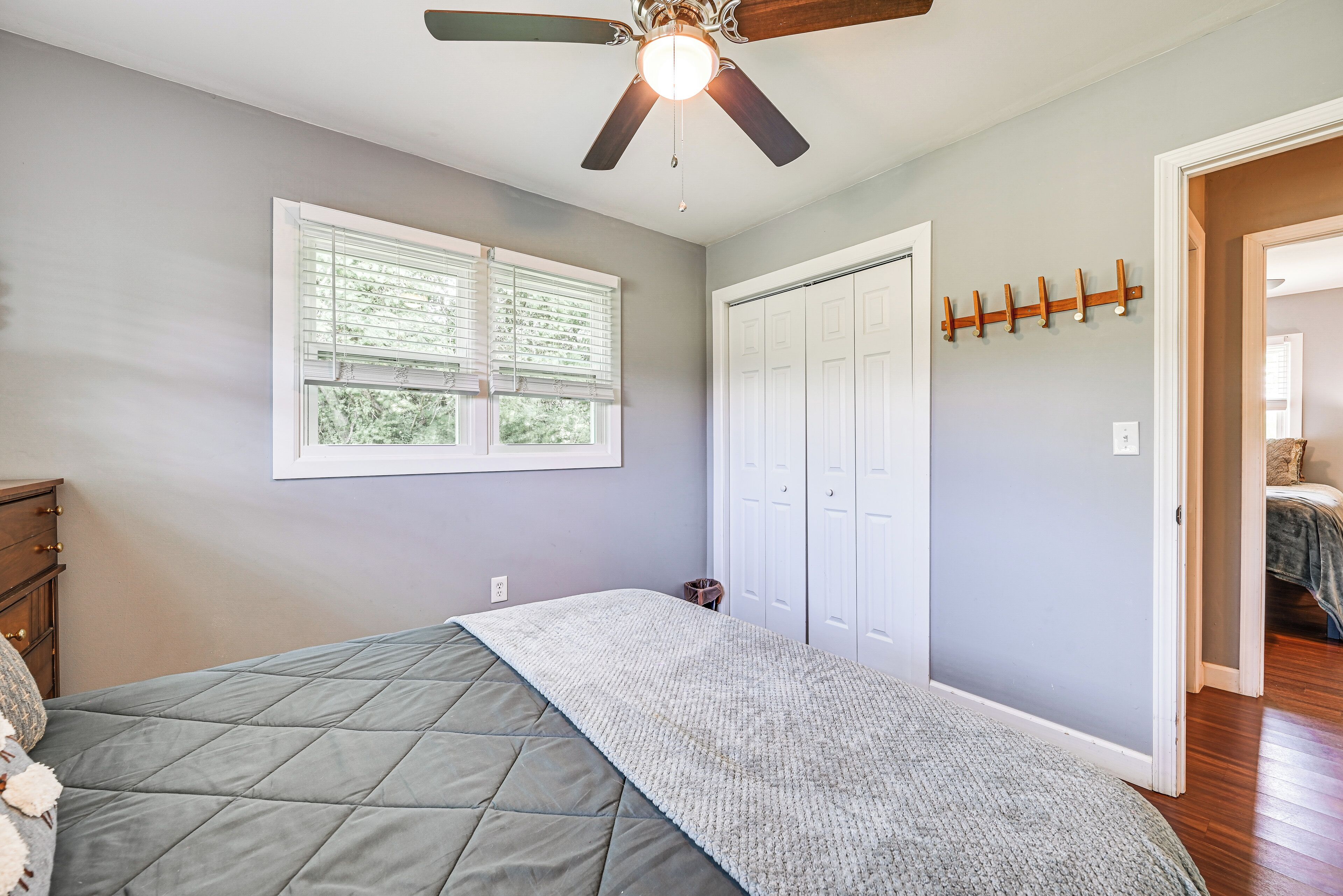 Un dormitorio con una cama, ventilador de techo, ventana con persianas, perchero y puerta a otra habitación.