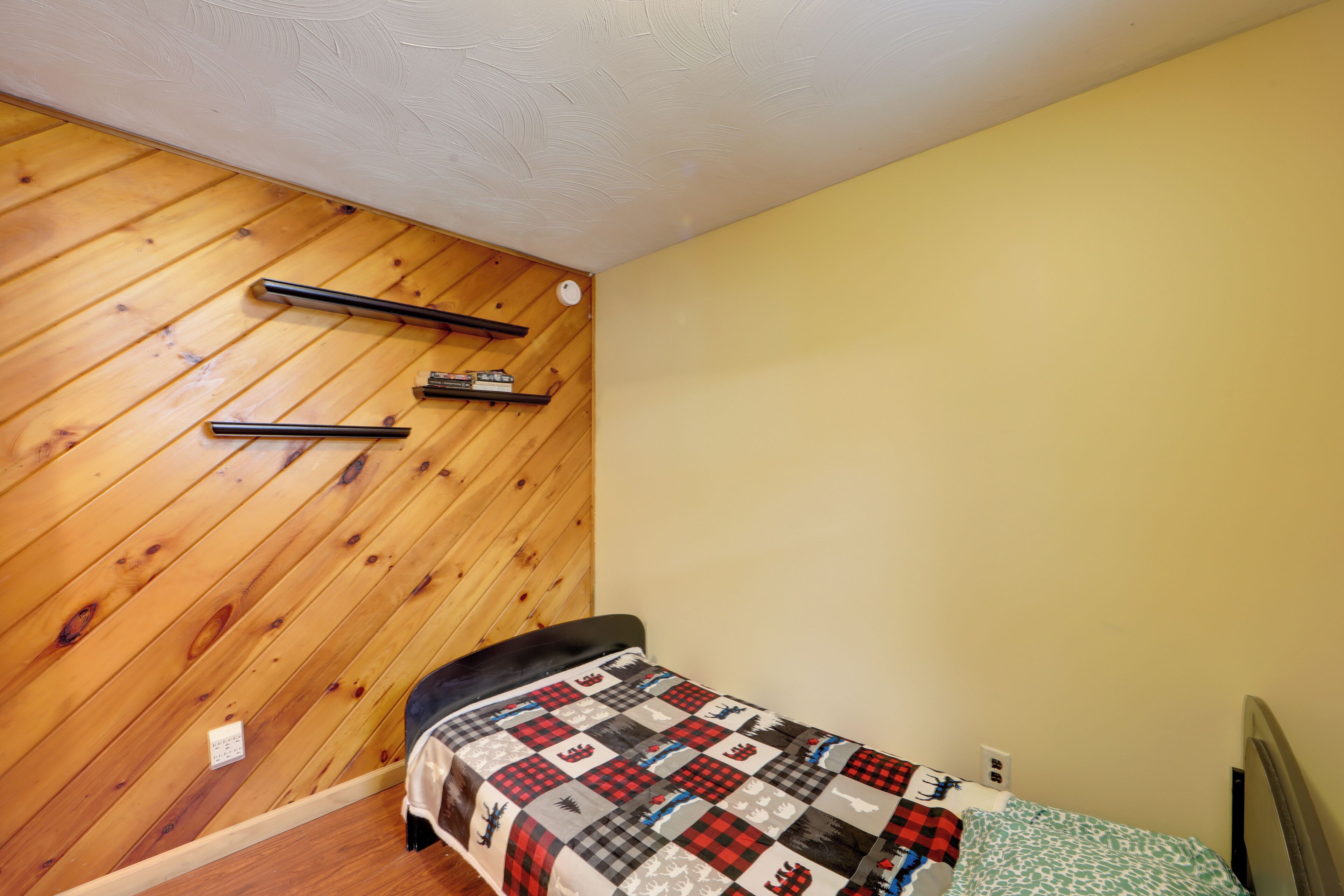 Un dormitorio pequeño con techo de listones de madera, una cama con colcha a cuadros y una mesita de noche.