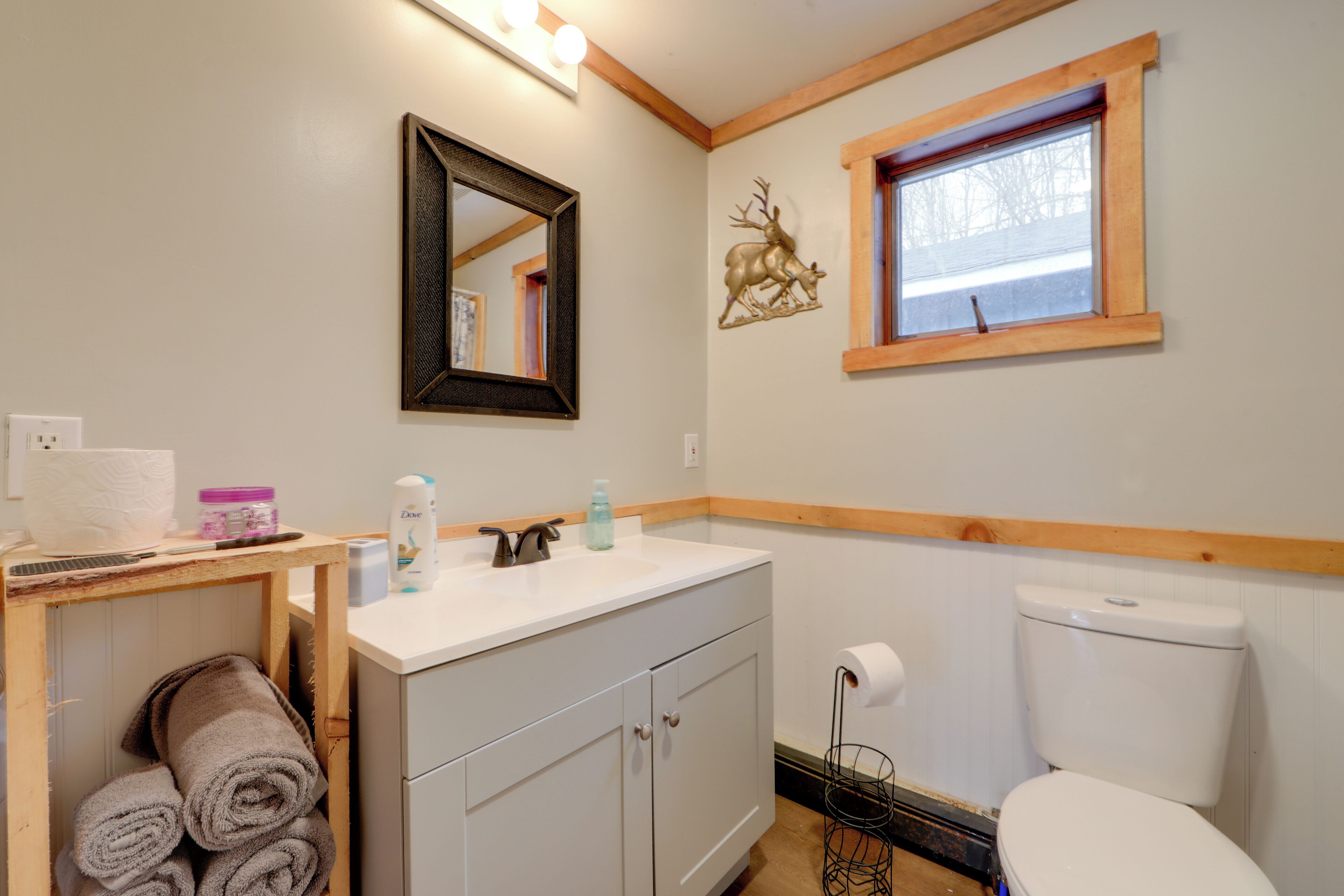 Un baño con lavamanos, espejo y retrete blancos. Hay toallas en un estante y una decoración de ciervo en la pared.