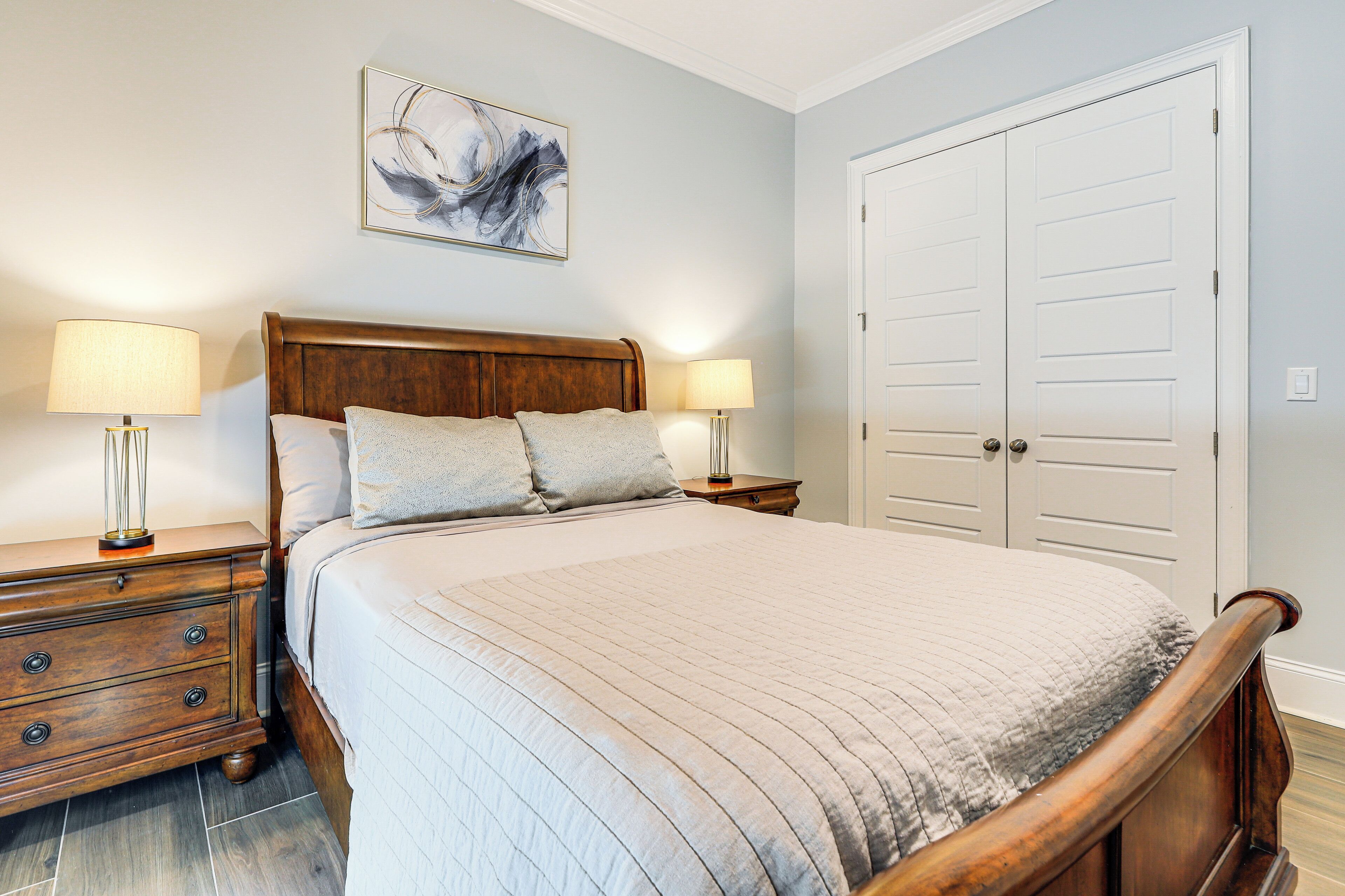 A bedroom with a wooden bed, two bedside tables with lamps, a large white door, and a wall-mounted abstract painting.