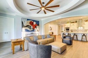 Un salon moderne avec un canapé, un pouf et une table basse. On y trouve également un coin cuisine avec des tabourets de bar et un ventilateur de plafond.