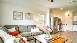 A living room with a grey sofa, a wooden coffee table, and a kitchen area with white cabinets and a stainless steel refrigerator. - 12 Mi to Avery Island: Spacious Family Home! (New Iberia)