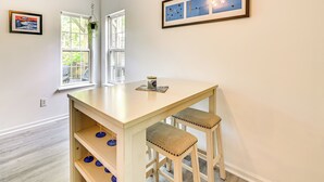 Een kamer met een lichte houten tafel, twee gestoffeerde krukjes, een hangplant en twee kunstwerken in lijsten aan de muur.