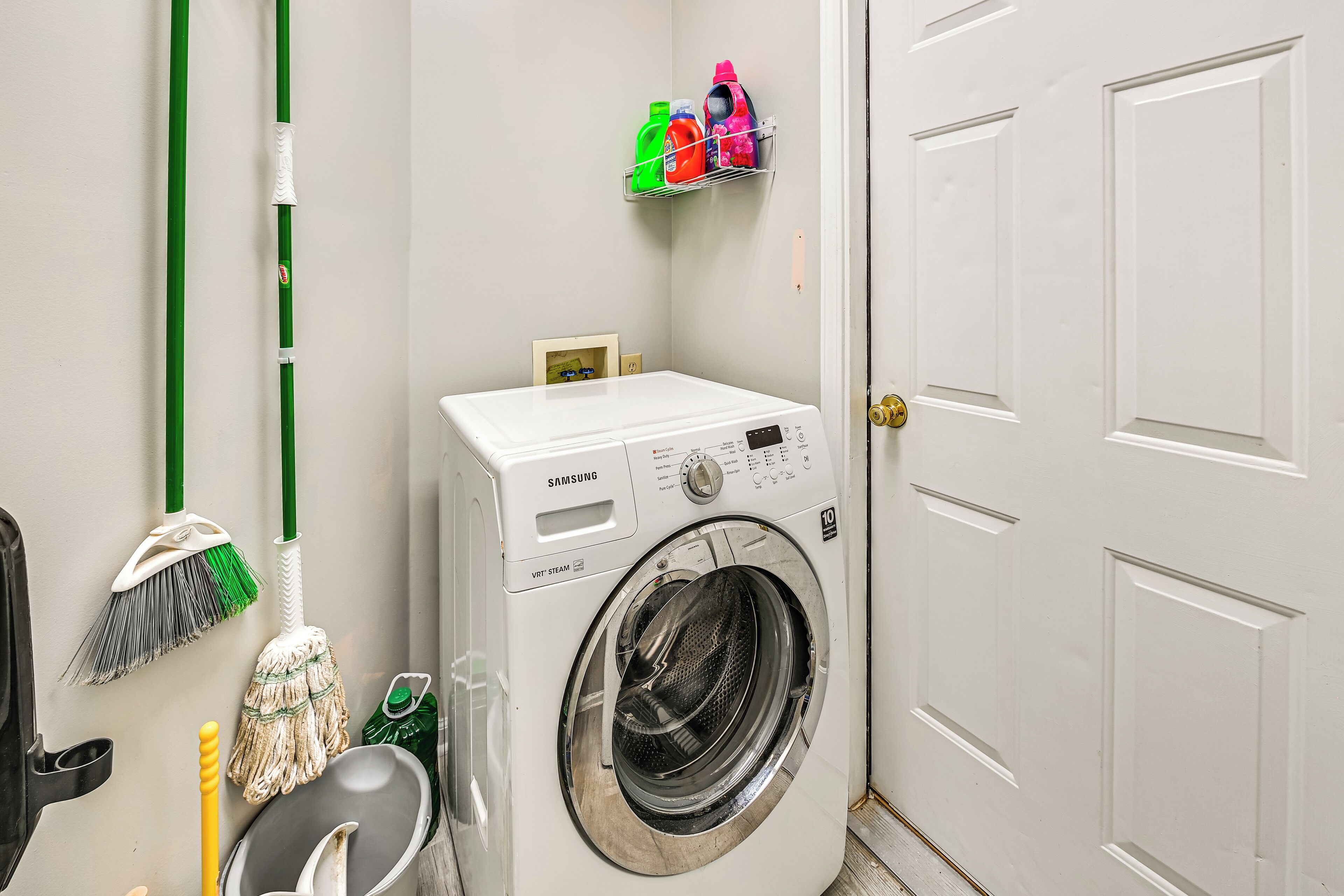 A laundry room with a Samsung washer and dryer, a mop, a broom, and a shelf with cleaning supplies.