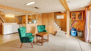 Een ruime kamer met houten vloer, een groene fauteuil, een houten salontafel, een keukenhoek met een wasmachine, en een trap naar een ander niveau.
