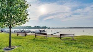 A grassy park area with benches and a tree, overlooking a calm body of water with boats docked in the background.