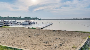 A sandy beach with a dock extending into calm water, surrounded by trees and boats.