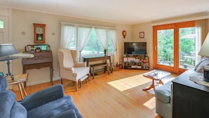 A living room with a wooden floor, a sofa, a chair, a desk, a television, and a window with curtains. - 1 Mi to Main St: Charming Elsworth Home w/ Yard! (Ellsworth)