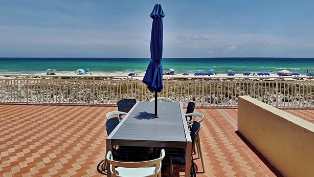 Une terrasse avec une table et des chaises, donnant sur une plage parsemée de parasols et sur l’océan.