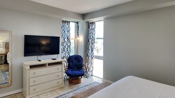A bedroom with a bed, a TV, a dresser, a chair, and a ceiling fan. - Sans Souci 102 | Gulf-Front, Large Patio + Seasonal Beach Service (Pensacola Beach)