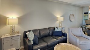 A living room with a black leather sofa, two side tables with lamps, a light-colored armchair, and a wall-mounted decorative plate. - Sans Souci 102 | Gulf-Front, Large Patio + Seasonal Beach Service (Pensacola Beach)