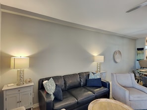 A living room with a black leather sofa, two side tables with lamps, a light-colored armchair, and a wall-mounted decorative plate. - Sans Souci 102 | Gulf-Front, Large Patio + Seasonal Beach Service (Pensacola Beach)