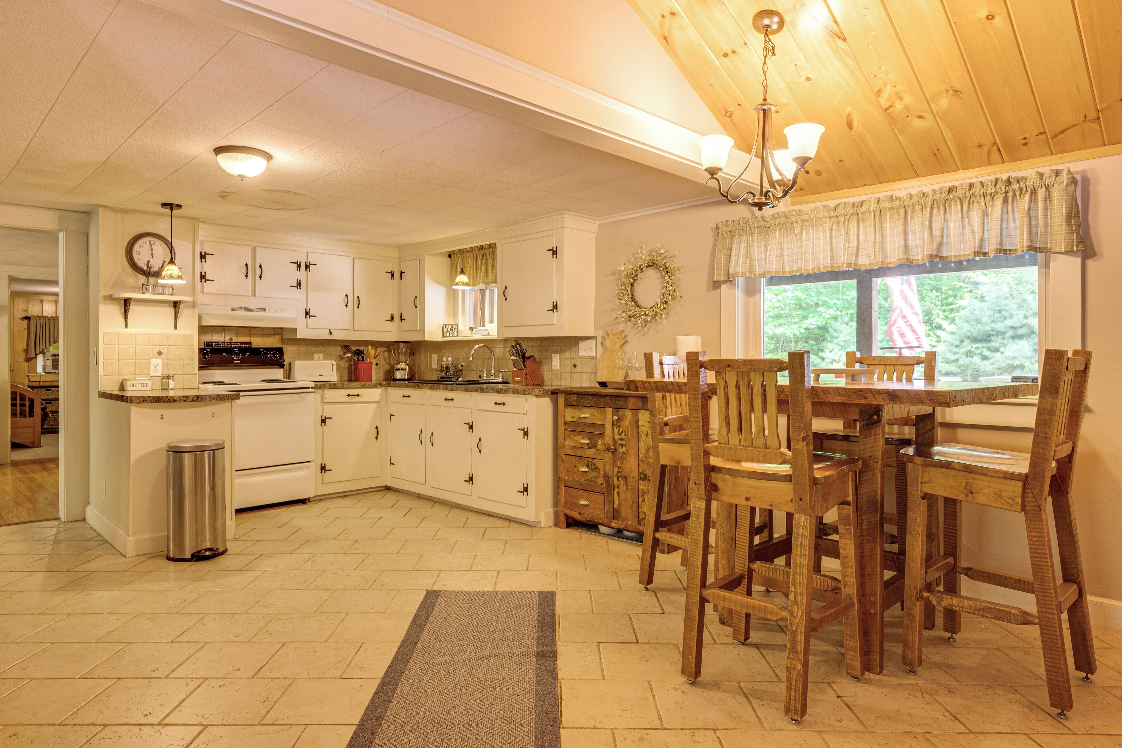 A kitchen with white cabinets, a wooden dining table with chairs, a trash can, and a window with a view of greenery.