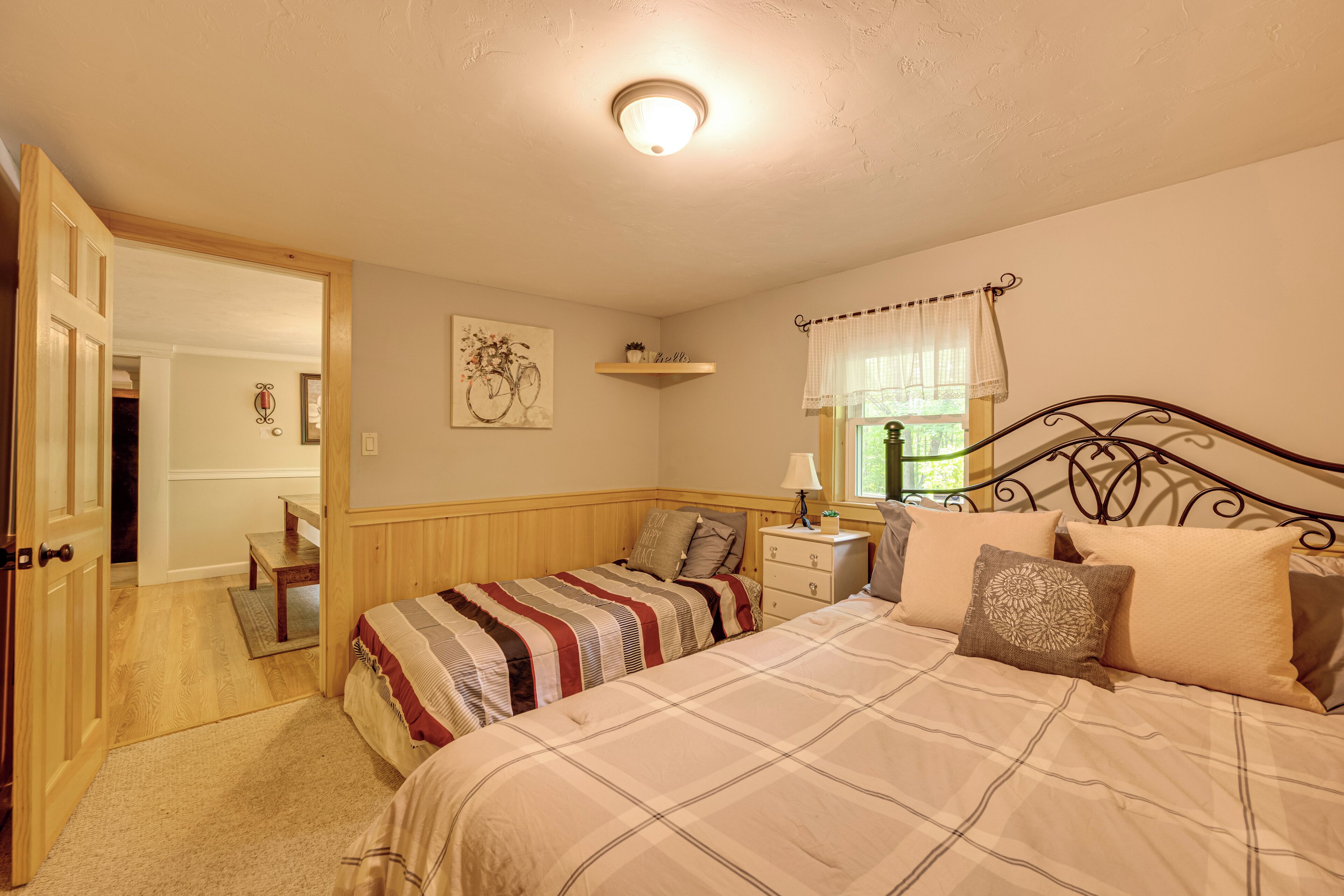 A bedroom with two beds, a nightstand, a dresser, a window with a curtain, and a door leading to another room.