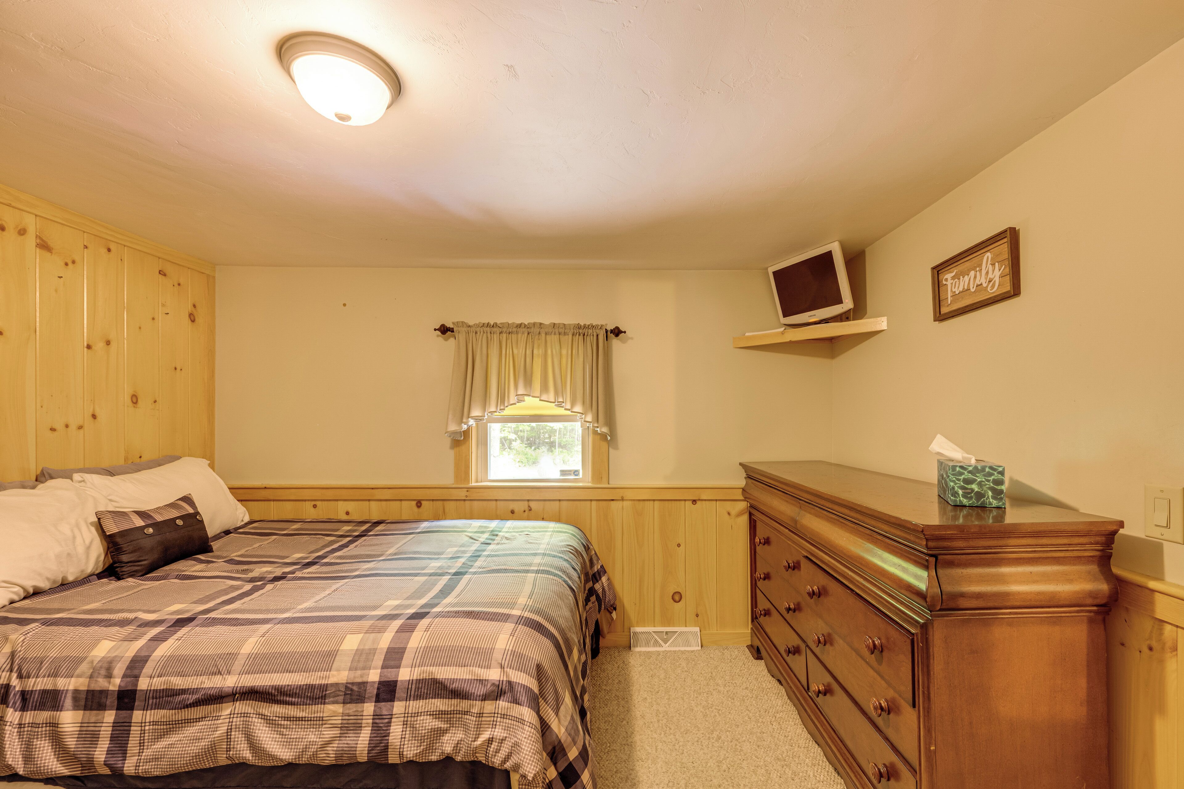 A bedroom with a bed, wooden dresser, a window with curtains, a television on a shelf, and a 'Family' sign on the wall.