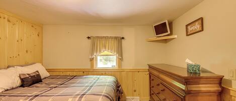 A bedroom with a bed, wooden dresser, a window with curtains, a television on a shelf, and a 'Family' sign on the wall.