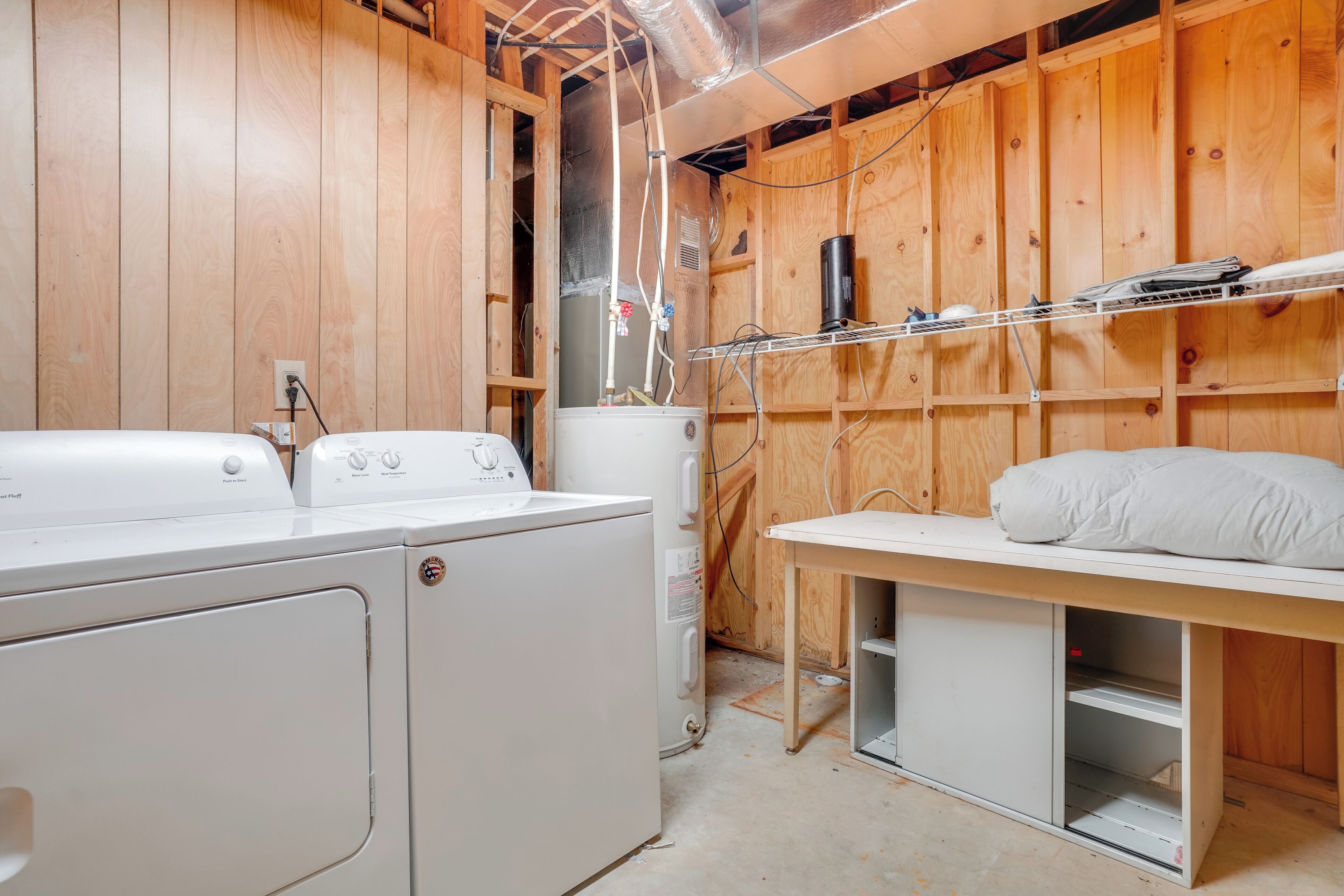 Un espace buanderie comprenant deux machines à laver blanches, un chauffe-eau et une étagère en bois sur laquelle est posé un coussin.