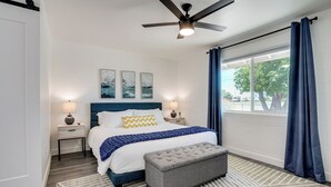 Une chambre à coucher comprenant un lit, des tables de chevet, un ventilateur de plafond et une grande fenêtre avec des rideaux.