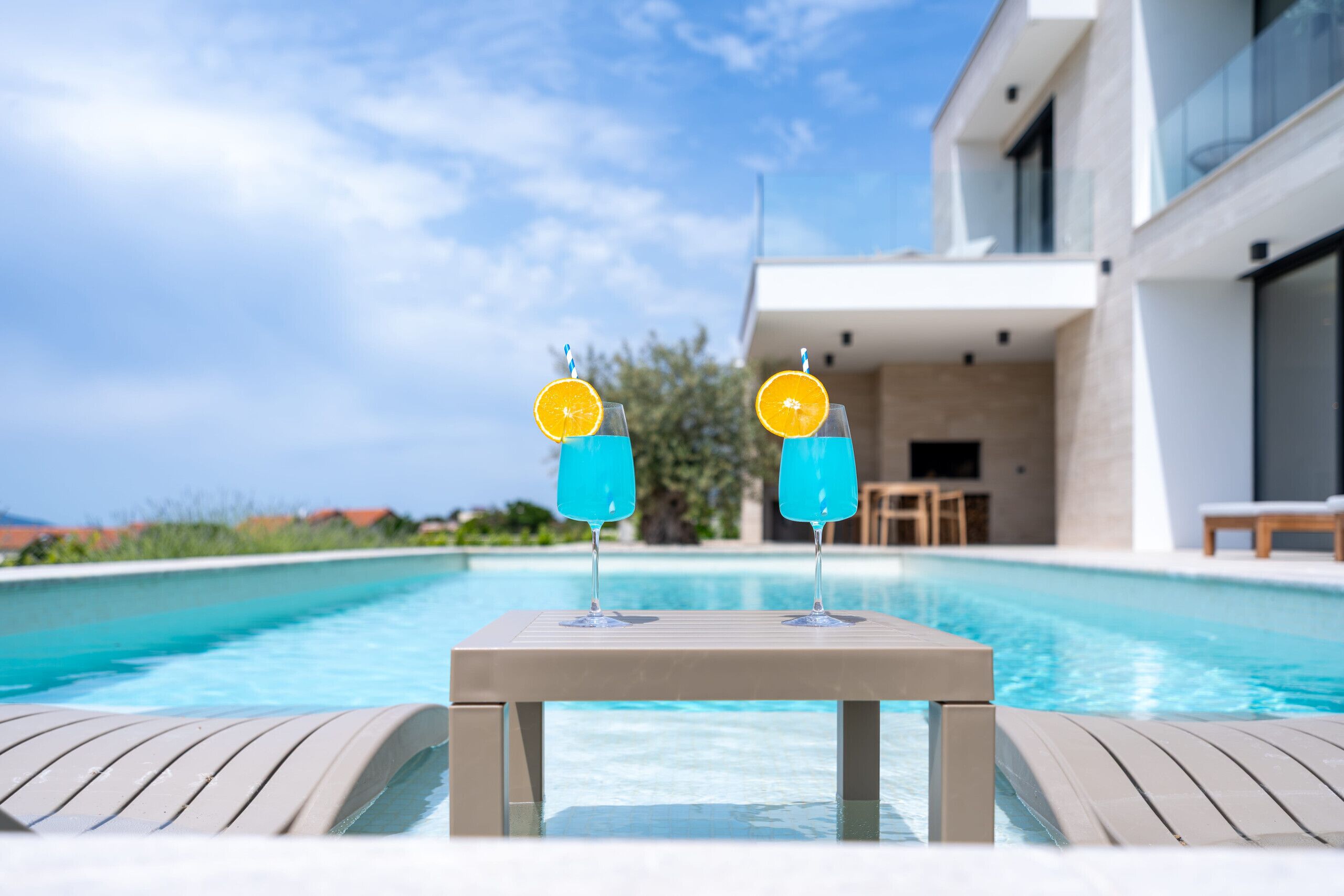 Una mesa junto a la piscina con dos vasos de un cóctel azul, un edificio moderno y un cielo despejado.