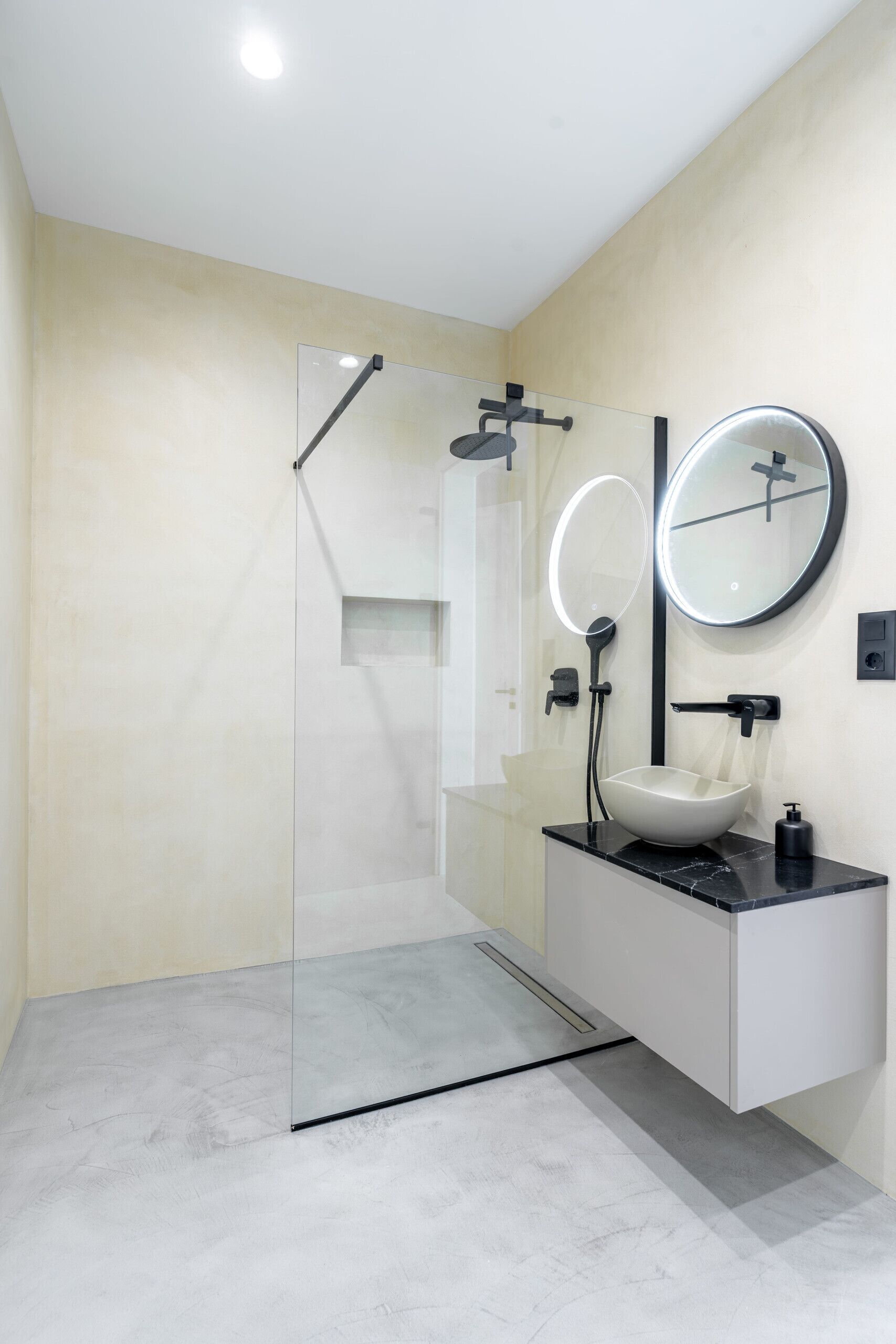 Un baño moderno con cabina de ducha de vidrio, un lavamanos blanco con encimera negra y un espejo redondo con luz superior.