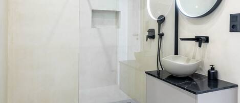 Un baño moderno con cabina de ducha de vidrio, un lavamanos blanco con encimera negra y un espejo redondo con luz superior.