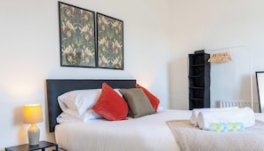 Ein ordentlich bezogenes Bett mit Kissen, einer Nachttischlampe, einem an der Wand montierten Regal mit Kleidung und zwei gerahmten Kunstwerken darüber.