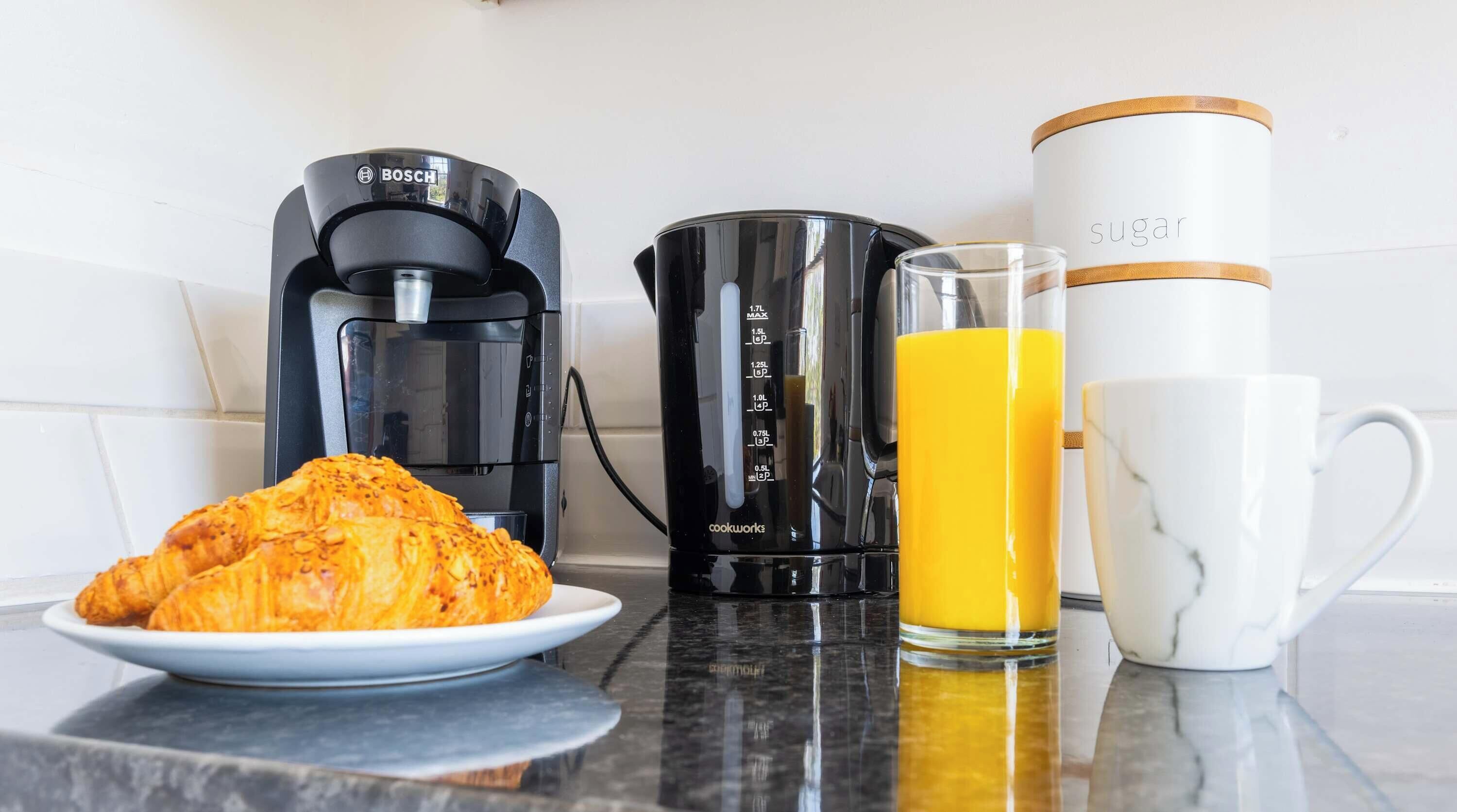 En køkkenbordplade med kaffemaskine, blender, et glas appelsinjuice, en hvid kop og en tallerken med croissanter.