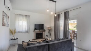 A modern living room with a grey sofa, a fireplace, a TV, and a dining area visible through a glass door. - Casa Platoni (Apolakkia)
