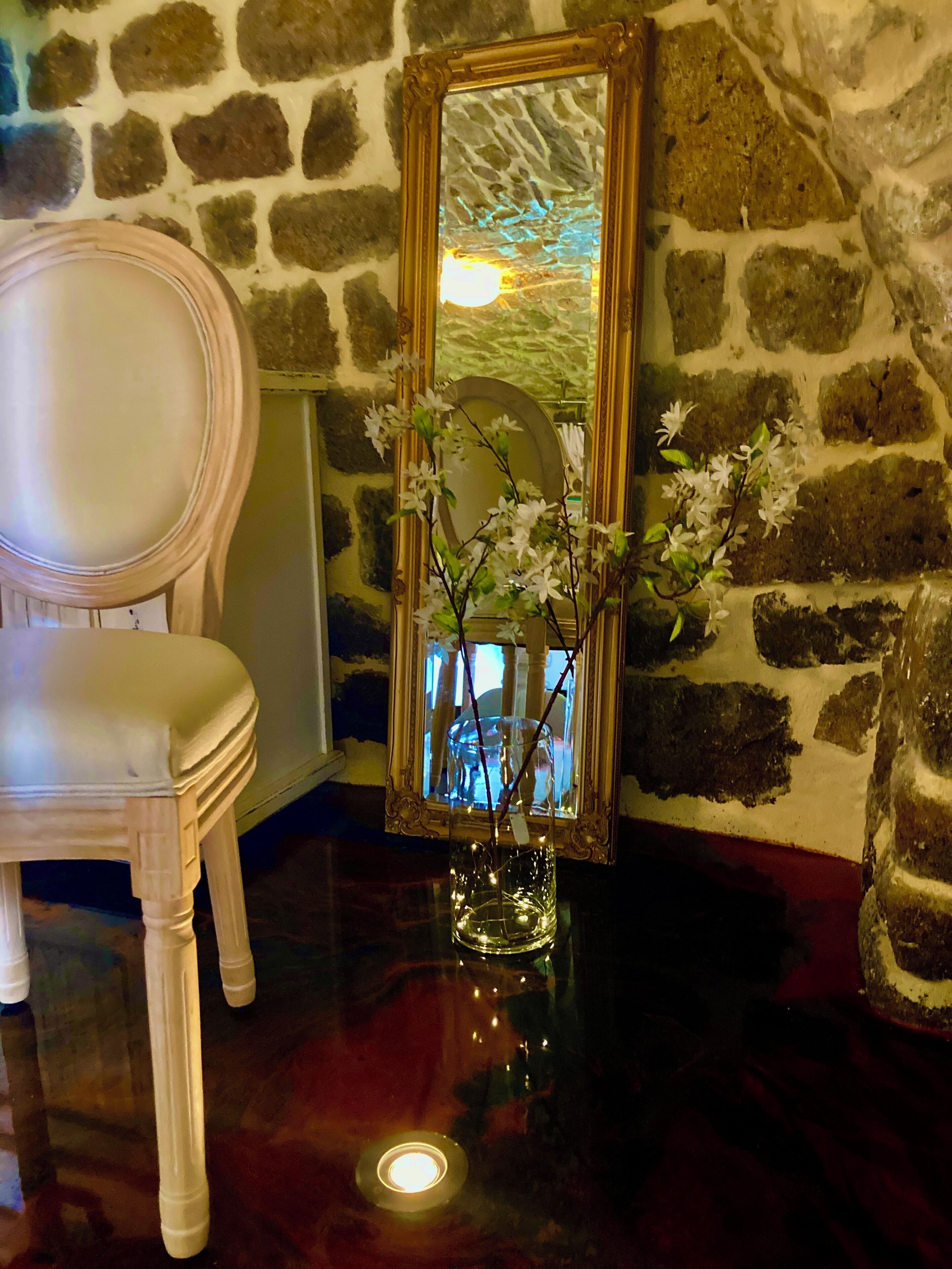 A room with a stone wall, a wooden chair, and a mirror with a vase of flowers.