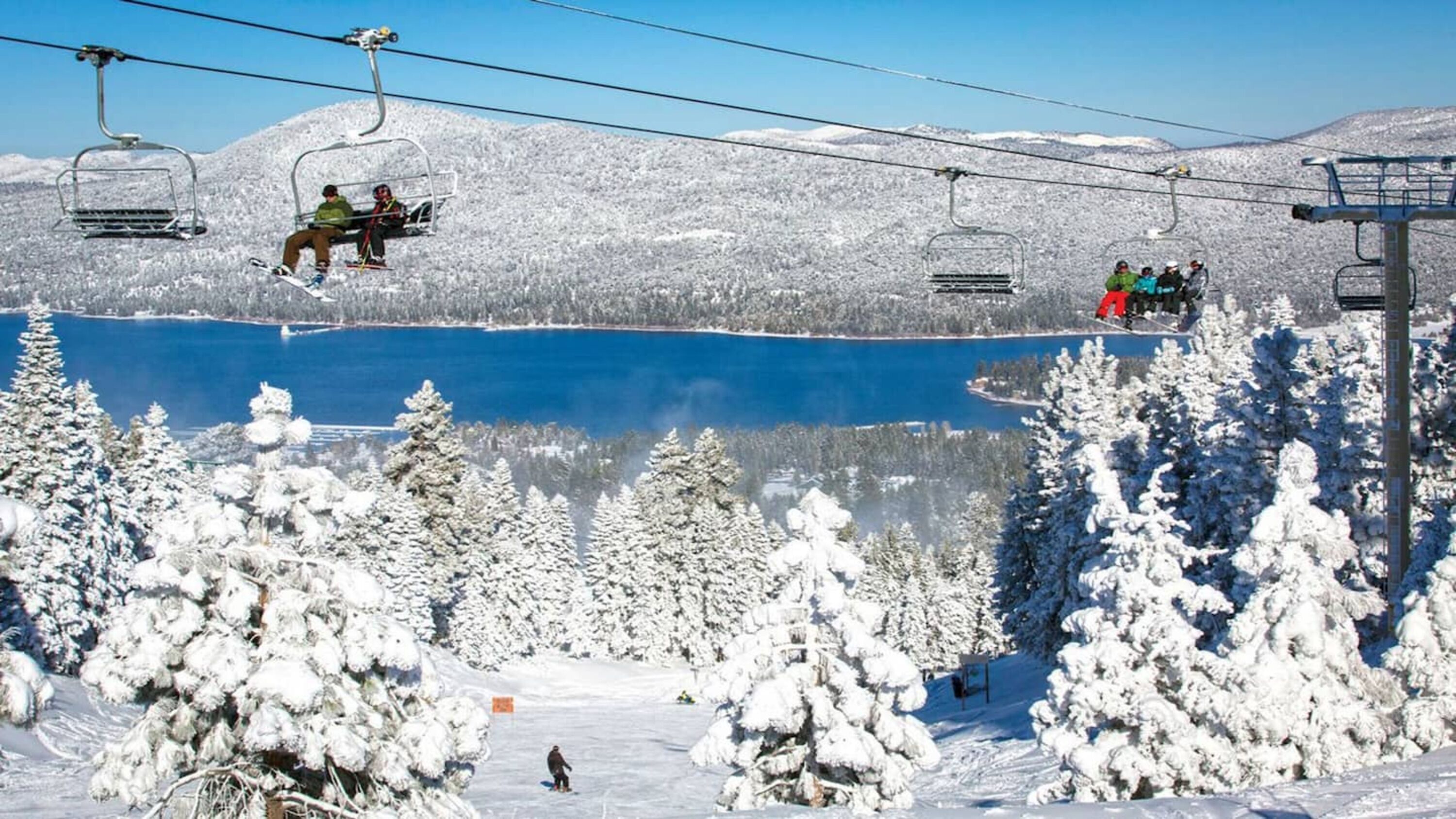 A ski lift with skiers and snowboarders, surrounded by snow-covered trees and a clear blue lake.