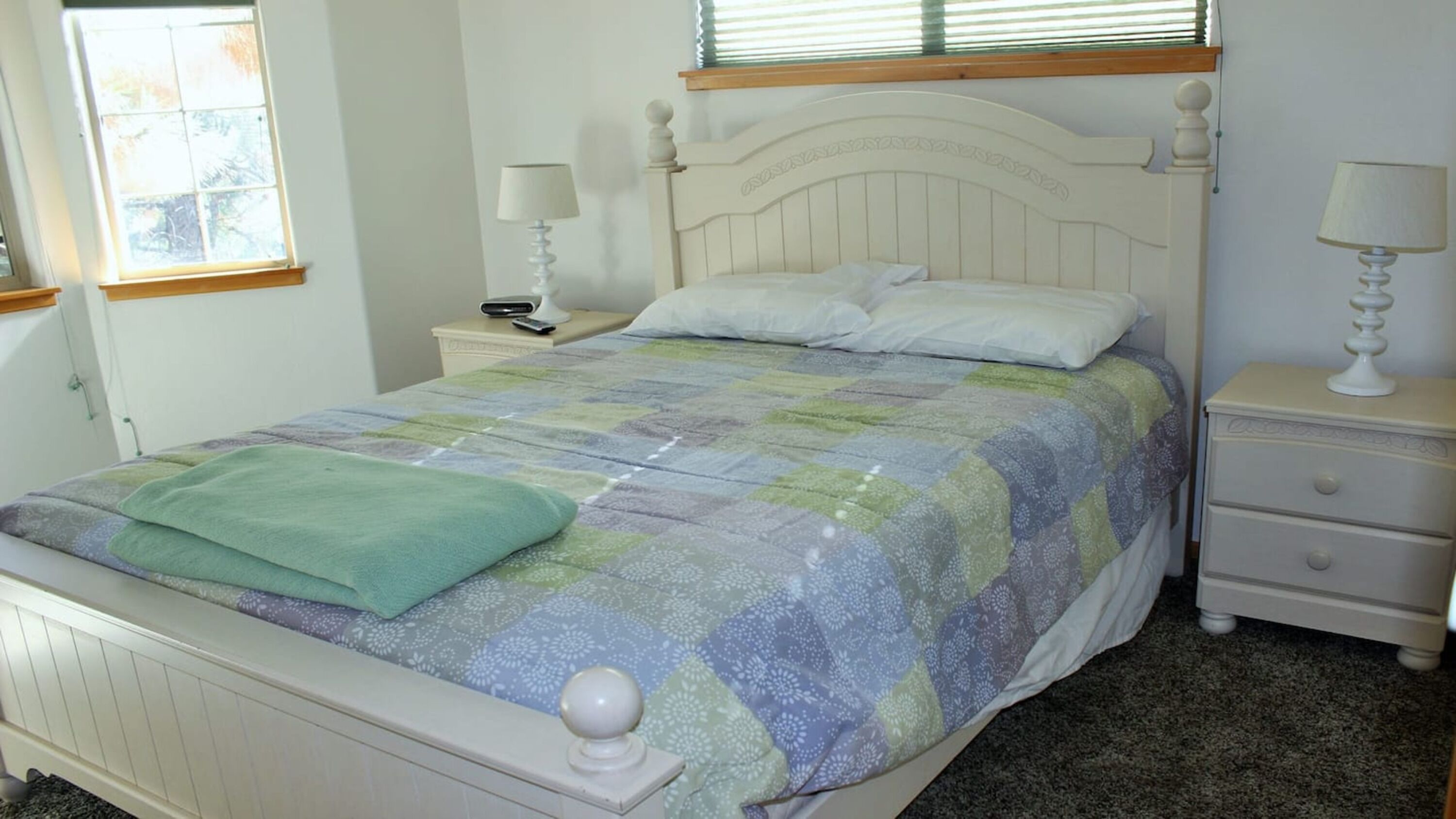 Una cama bien tendida con un edredón estampado, dos mesitas de noche con lámparas y una ventana con persianas.