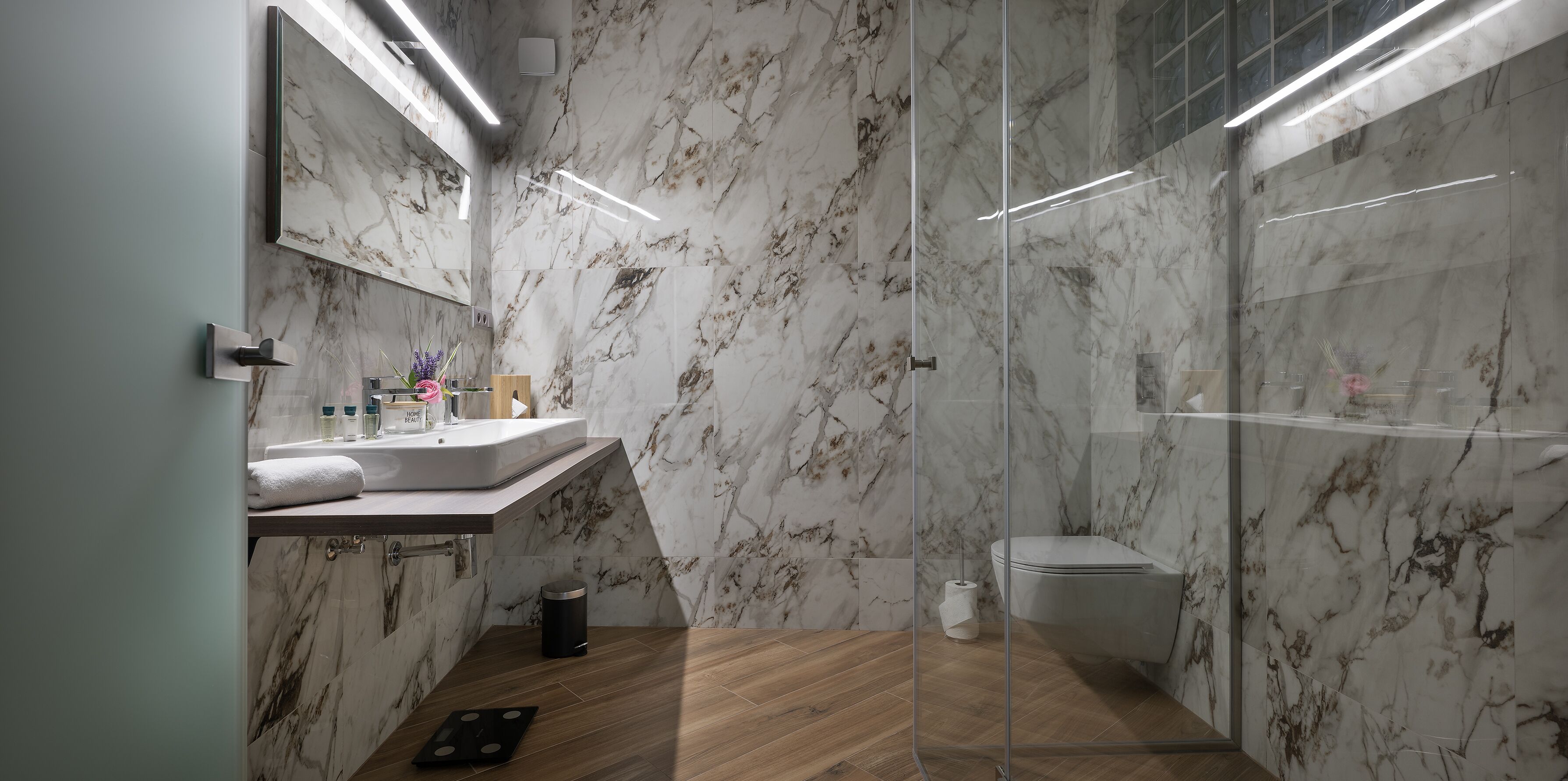 Ein modernes Badezimmer mit Marmorwänden, Holzboden, einer Glasdusche und einem Waschtisch mit Spiegel und Waschbecken.