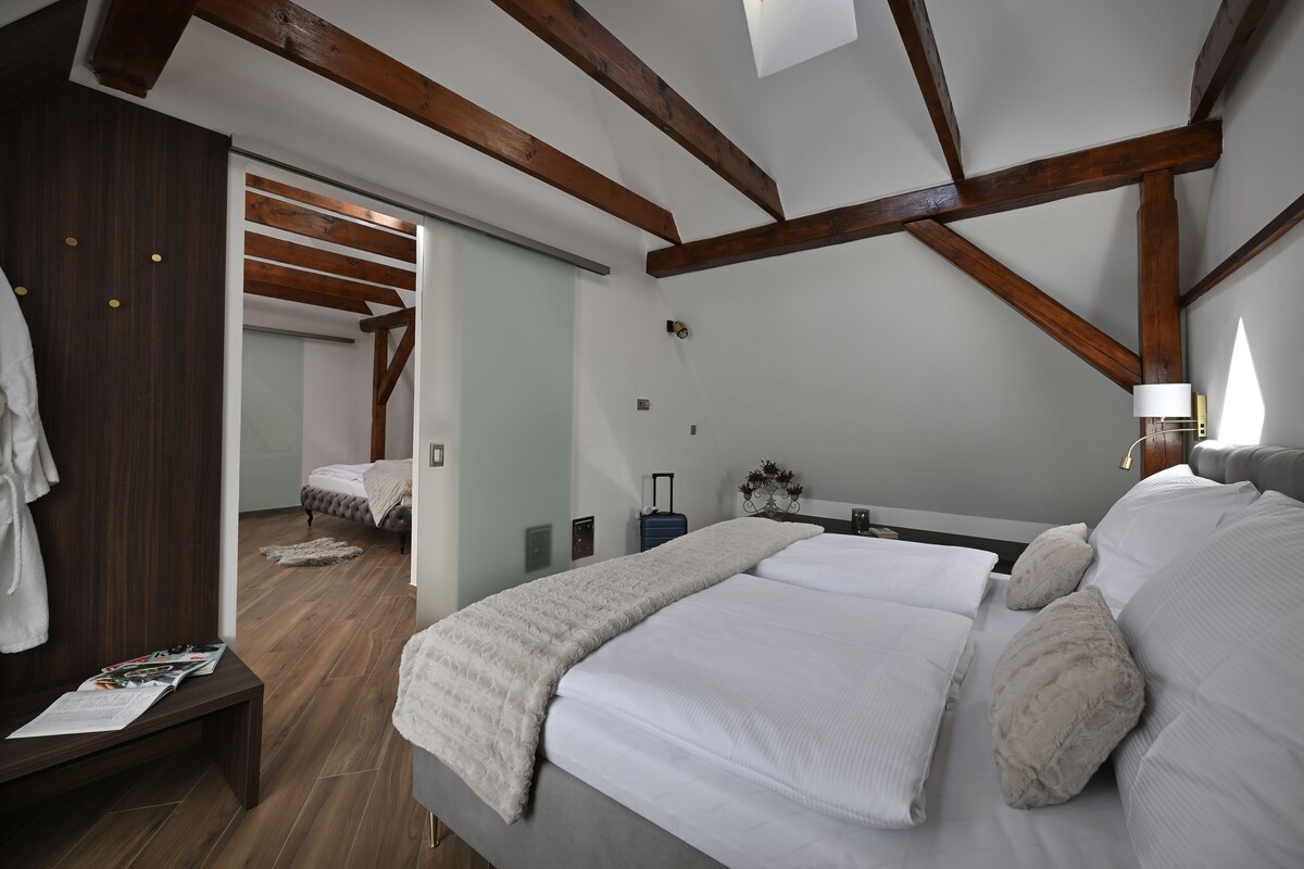 A bedroom with a large bed, wooden beams, a bench, a suitcase, and a nightstand with a lamp.