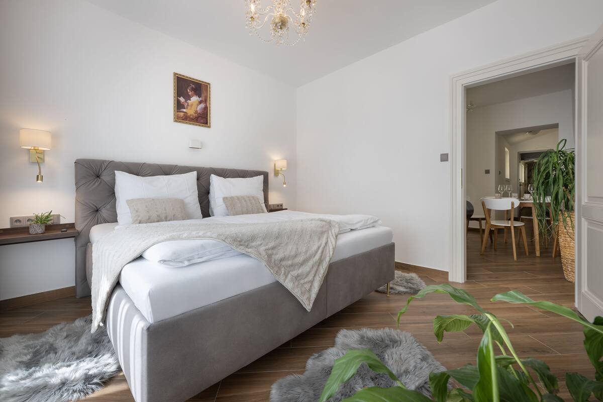 A modern bedroom with a large bed, bedside tables, a chandelier, and a dining area visible through an open door.