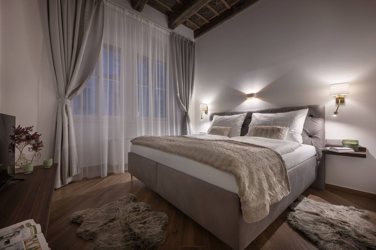 A bedroom with a large bed, wooden floor, fur rugs, a nightstand, and wall-mounted lamps.