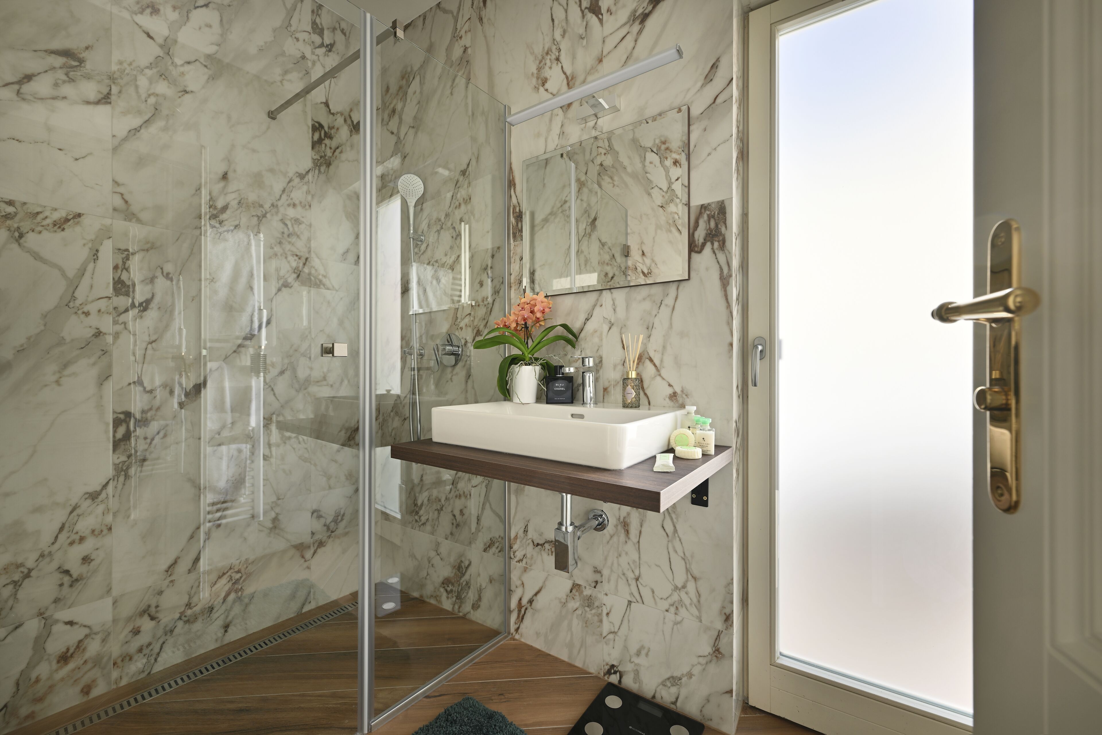 Un baño con pared de mármol, cabina de ducha de vidrio, lavamanos con espejo y puerta con pomo de latón.