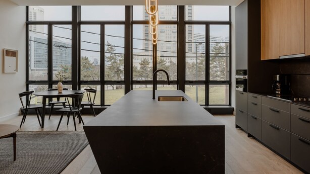A modern kitchen with a large island, dark cabinetry, and a view of a cityscape through large windows. - Sosuite One Bed Apt Accessible w Rooftop Gym Laundry (Philadelphia)