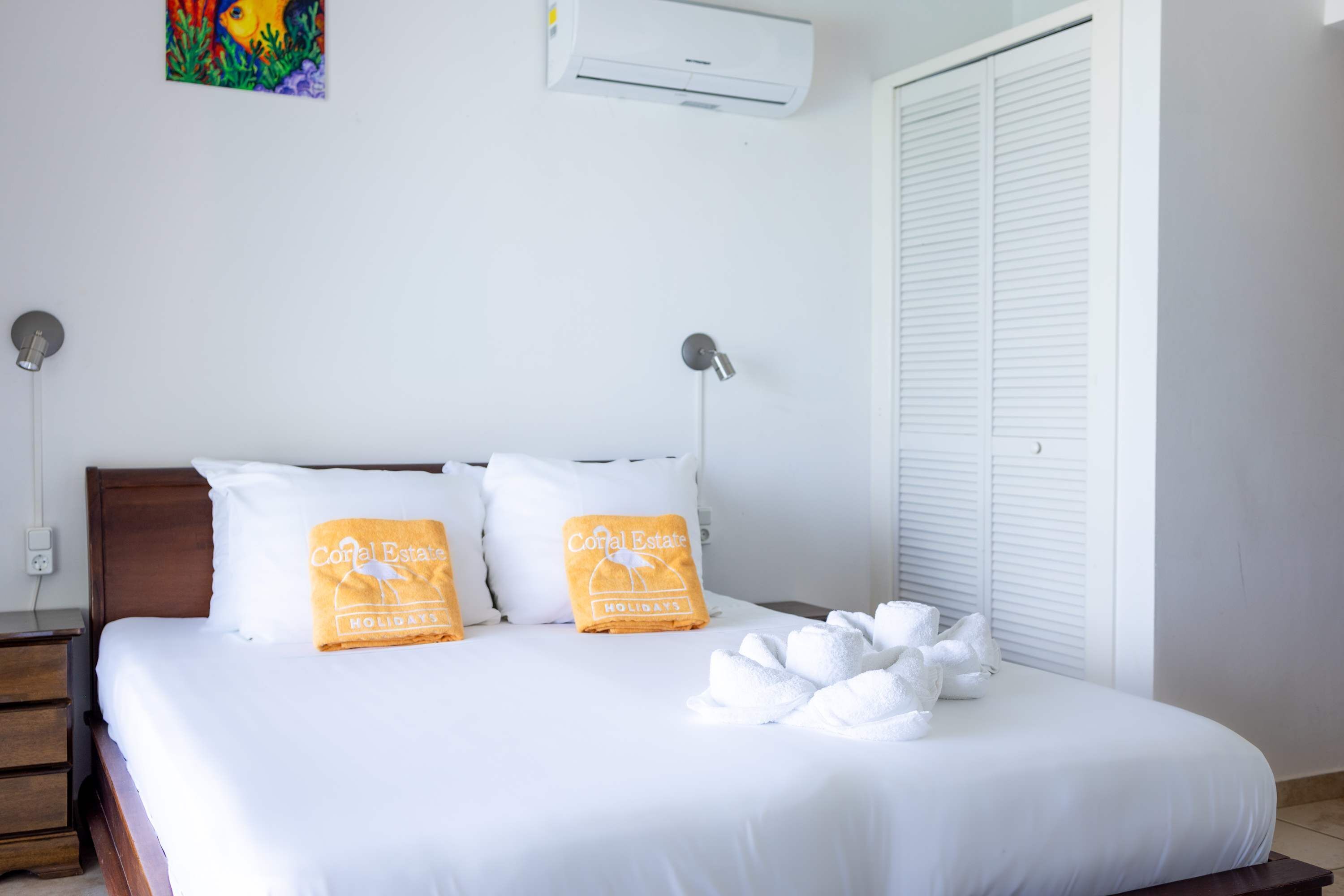 En pænt redt seng med hvide sengetøj og to orange puder, en sengetårnslampe samt en hvid aircondition-enhed på væggen.