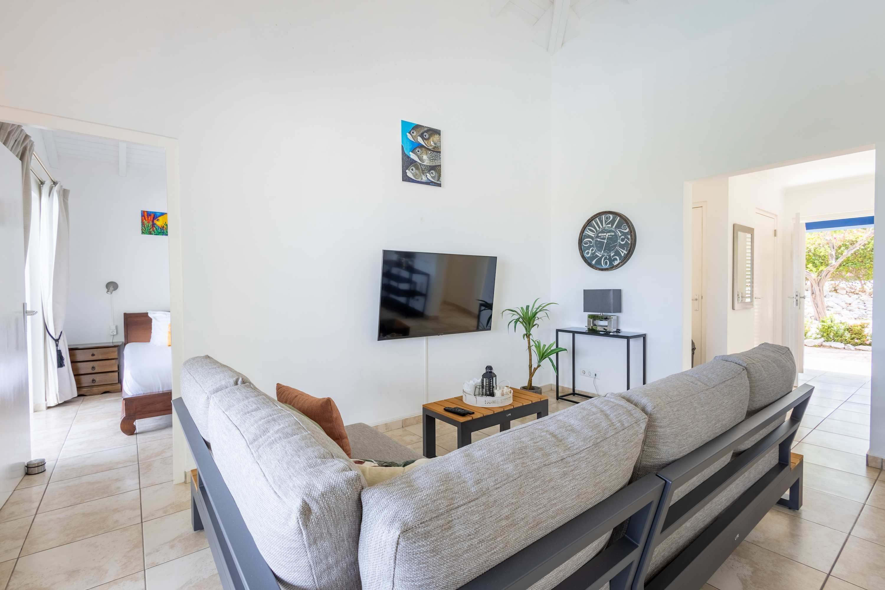 A modern living room with a large sofa, a flat-screen TV, a small table with a plant, and a clock on the wall.