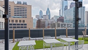 A rooftop terrace with white metal tables and chairs, a small patch of artificial grass, and a city skyline view in the background. - Sosuite One Bed Apt Plus w Rooftop, Gym, Laundry (Philadelphia)