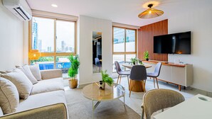 Een moderne woonkamer met een bank, een eettafel, stoelen, een televisie en grote ramen met uitzicht op de stad.