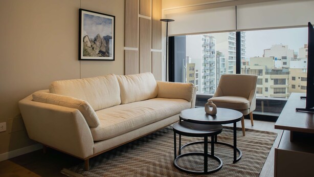Ein modernes Wohnzimmer mit einem beigen Sofa, einem runden Couchtisch und einem Sessel. An der Wand hängt ein gerahmtes Bild und ein großes Fenster bietet Blick auf die Stadt.