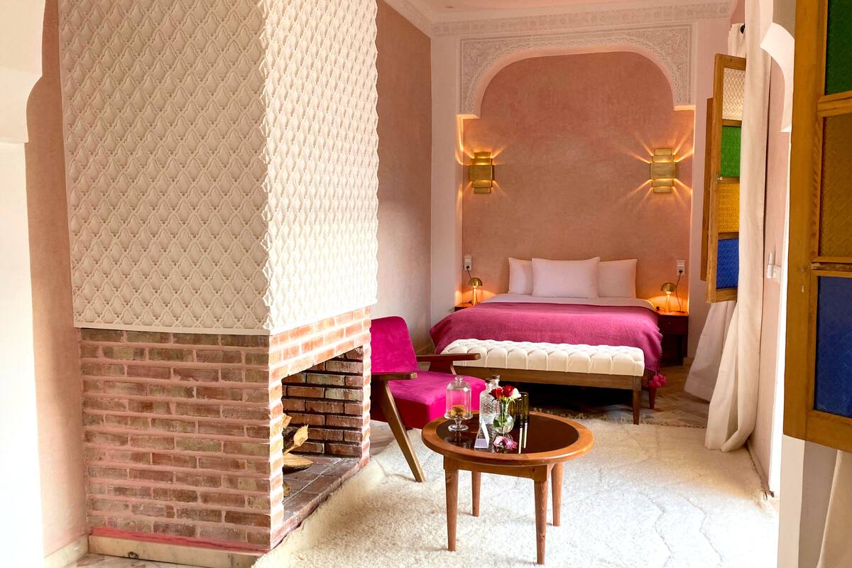 Een knus slaapkamer met een schouw van baksteen, een bed met een roze sprei, twee wandlampen en een klein houten tafeltje met een vaas.