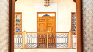 Une porte en bois traditionnelle, agrémentée d’un portail en métal décoratif, se dresse devant un mur de couleur beige orné de motifs métalliques complexes sur les fenêtres.
