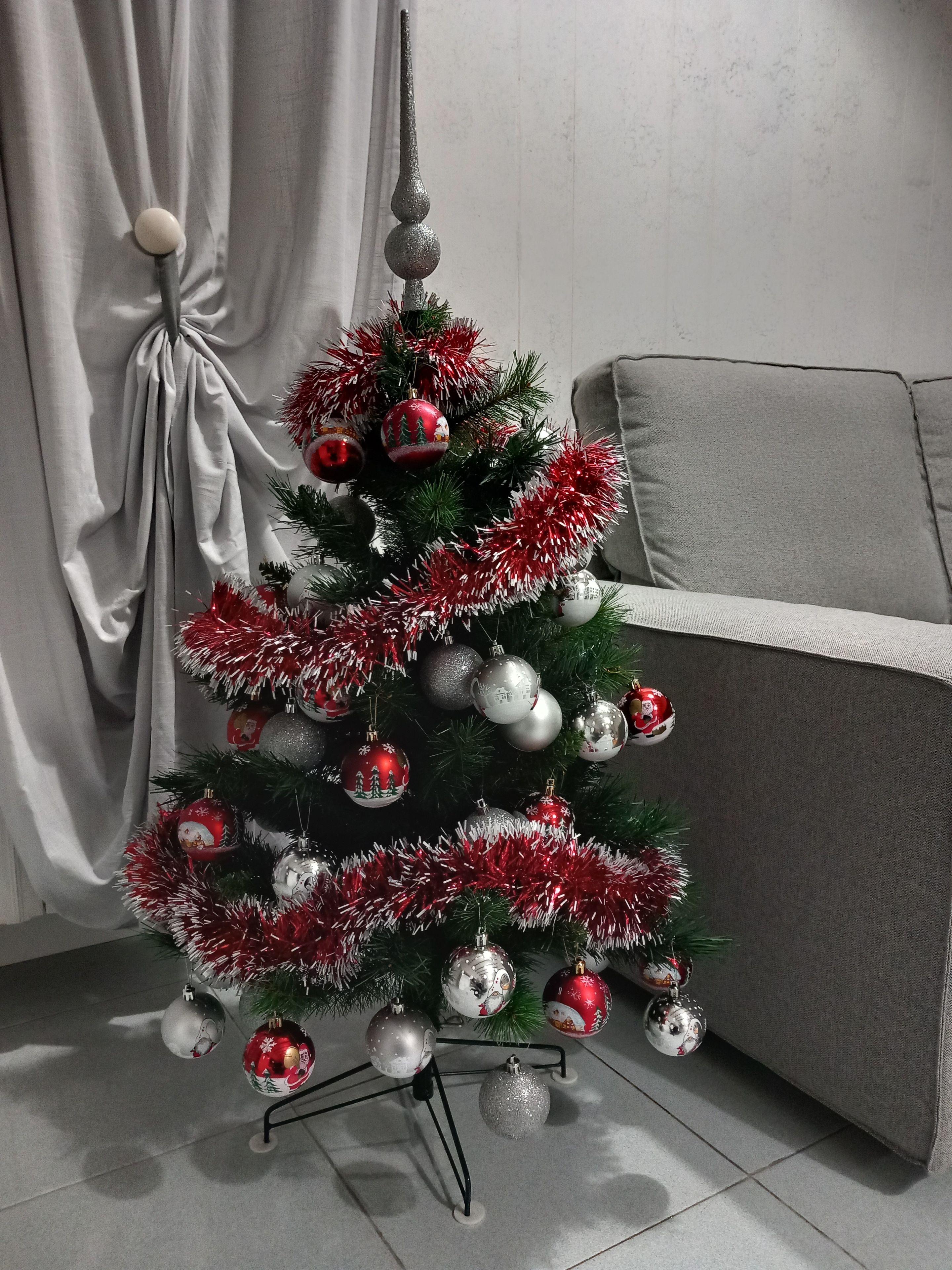 Árvore de Natal com enfeites e guirlanda, em um quarto com sofá cinza e cortinas.