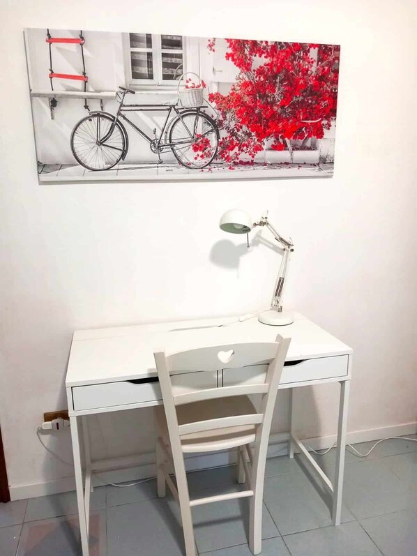 Un bureau blanc avec une chaise, une lampe de bureau et un tableau représentant un vélo et des fleurs accroché au mur.