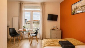A hotel room with a bed, a desk with a chair, a small table with a chair, a TV on the wall, and a balcony with a view of buildings. - Charmant studio terrasse en centre-ville (Châtellerault)