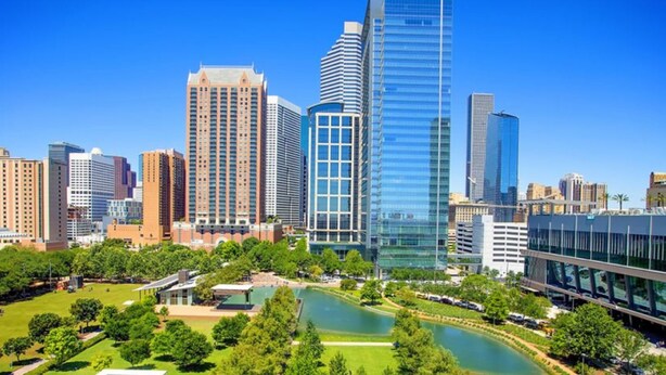 A cityscape with modern high-rise buildings, a central park with a pond, and a clear blue sky.